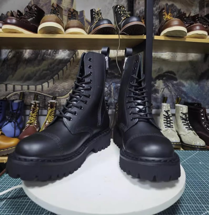 May include: Black leather combat boots with a lace-up design and side zippers. The boots have a thick, lugged sole and are displayed on a white surface. Other boots are visible on shelves in the background.