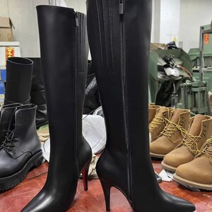 Puede incluir: Un par de botas negras hasta la rodilla con punta y tacones de aguja. Las botas tienen una cremallera lateral y est&aacute;n hechas de un material liso y brillante. Otras botas son visibles en el fondo.