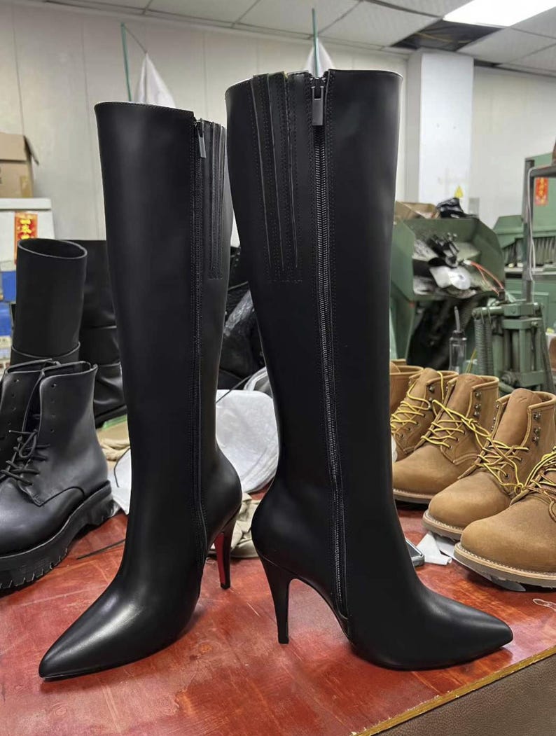 Puede incluir: Un par de botas negras hasta la rodilla con punta y tacones de aguja. Las botas tienen una cremallera lateral y est&aacute;n hechas de un material liso y brillante. Otras botas son visibles en el fondo.