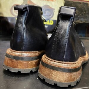 May include: Pair of black leather ankle boots with a thick, distressed brown sole and a black rubber tread. The boots have a pull tab at the back and a subtle, vintage aesthetic. The background features a motorcycle-themed poster.