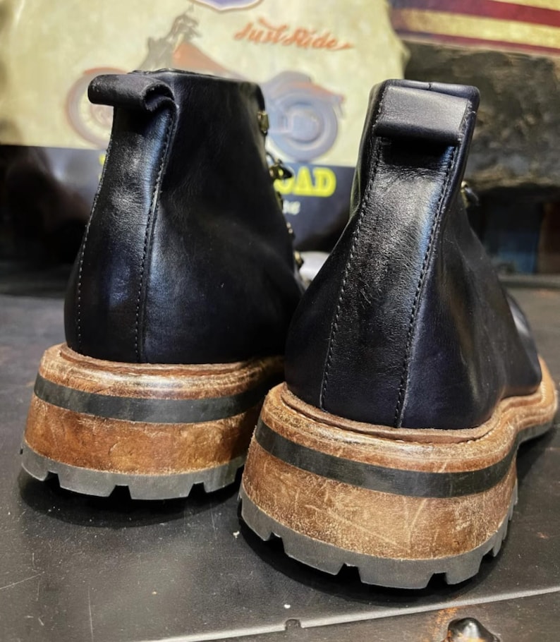May include: Pair of black leather ankle boots with a thick, distressed brown sole and a black rubber tread. The boots have a pull tab at the back and a subtle, vintage aesthetic. The background features a motorcycle-themed poster.