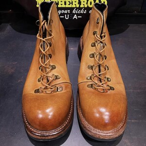 May include: A pair of light brown leather boots with a lace-up design and metal eyelets. The boots have a rounded toe and a thick, dark brown sole. The boots are displayed on a dark surface.