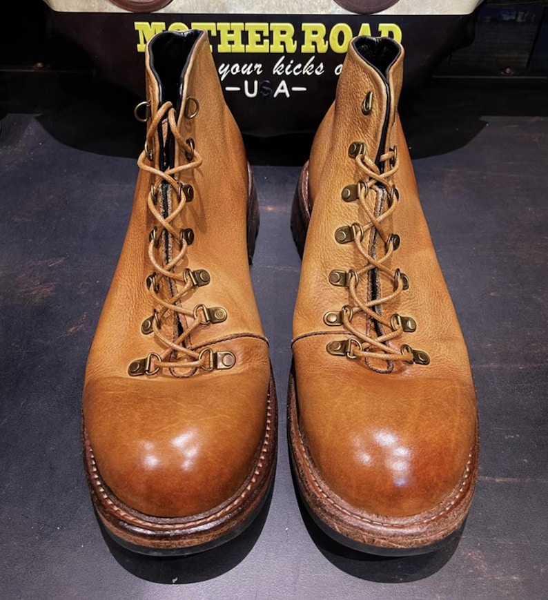 May include: A pair of light brown leather boots with a lace-up design and metal eyelets. The boots have a rounded toe and a thick, dark brown sole. The boots are displayed on a dark surface.