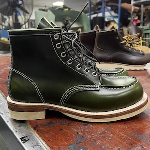 May include: Green leather work boot with white stitching and a light brown sole. The boot has a lace-up design with silver eyelets. Another brown boot is visible in the background.