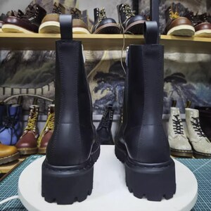 May include: Two black leather boots with thick, lug soles are displayed. The boots have a side zipper and a pull tab at the top. Other boots are visible on shelves in the background, showcasing a variety of styles and colors.