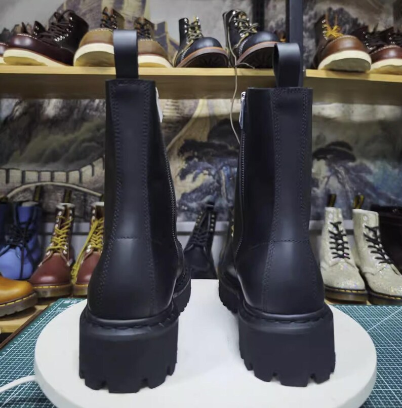 May include: Two black leather boots with thick, lug soles are displayed. The boots have a side zipper and a pull tab at the top. Other boots are visible on shelves in the background, showcasing a variety of styles and colors.