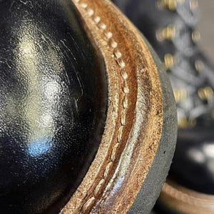 May include: Close-up of a black leather boot with a brown leather sole. The sole is stitched with a light-colored thread. The boot has a glossy finish and a rugged design, suggesting durability and quality craftsmanship.