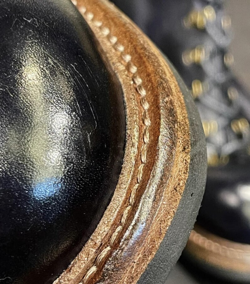 May include: Close-up of a black leather boot with a brown leather sole. The sole is stitched with a light-colored thread. The boot has a glossy finish and a rugged design, suggesting durability and quality craftsmanship.