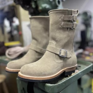 以下が含まれることがあります： 複数のバックル留めと丸いつま先が付いた、タン色のスエードブーツのペア。ブーツはミッドカーフ丈で、明るい茶色のエッジが付いた革底です。ブーツは緑色の表面に表示されています。ブーツの高さは約20cmです。