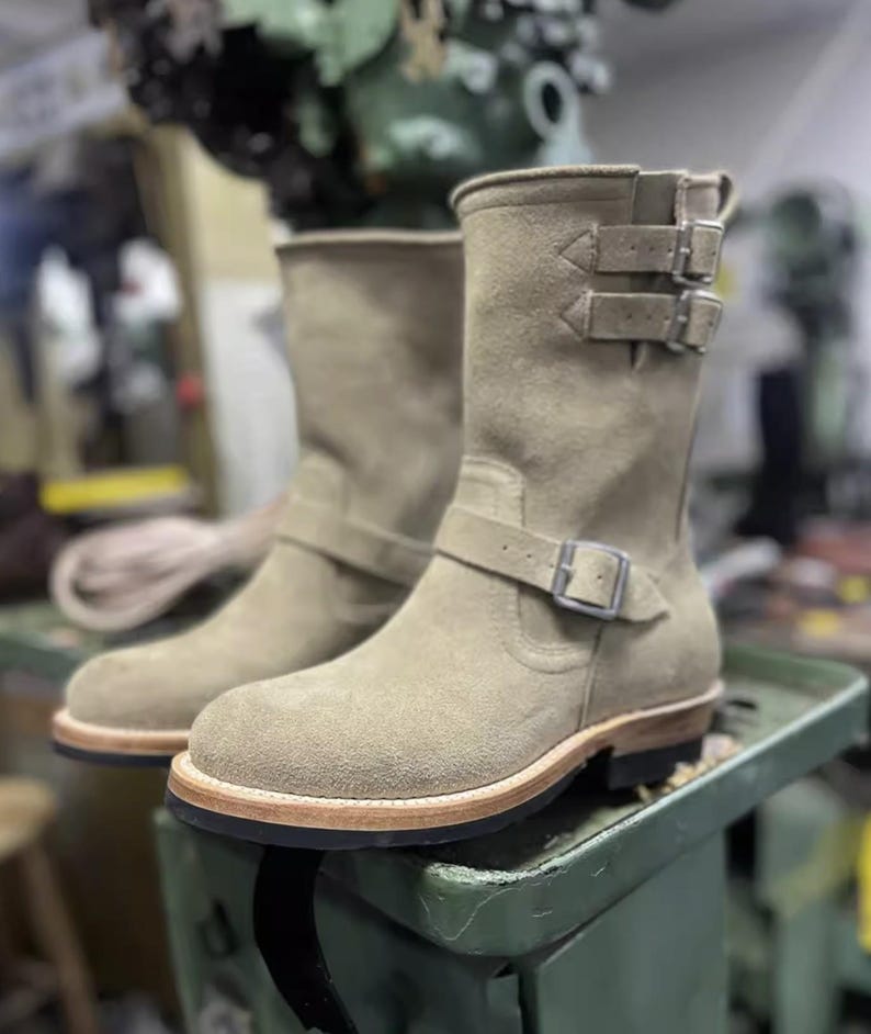 May include: Tan suede boots with buckle fastenings and a round toe. The mid-calf boots have a leather sole with a light brown edge. The boots are displayed on a green surface. The boots' height is approximately 20 cm.