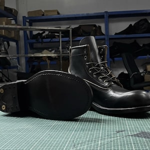 Op de afbeelding: Zwarte leren werklaarzen met een vetersluiting en een stevige zool. De laarzen hebben een klassieke stijl met zichtbare stiksels en metalen vetergaten. De zool is ongeveer 2,5 cm dik.