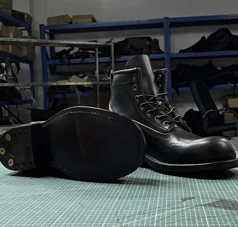 Op de afbeelding: Zwarte leren werklaarzen met een vetersluiting en een stevige zool. De laarzen hebben een klassieke stijl met zichtbare stiksels en metalen vetergaten. De zool is ongeveer 2,5 cm dik.