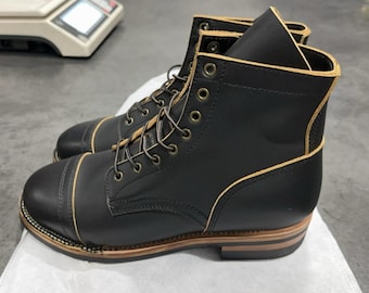 May include: A pair of black leather boots with cap toes and tan accents. The boots have a lace-up design with brass eyelets and a sturdy brown sole. The edges are trimmed with tan stitching.