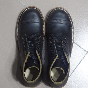 May include: A pair of black leather lace-up boots with a cap toe design. The boots have a brown leather sole and a green leather lining.