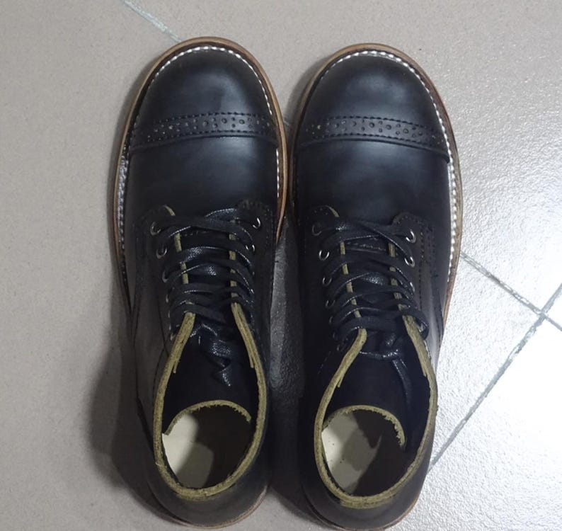 May include: A pair of black leather lace-up boots with a cap toe design. The boots have a brown leather sole and a green leather lining.