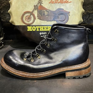 May include: Black leather boot with a rounded toe and brown sole. The boot features metal eyelets and a lace-up closure. The background includes a motorcycle graphic and the text "MOTHER your -U-".