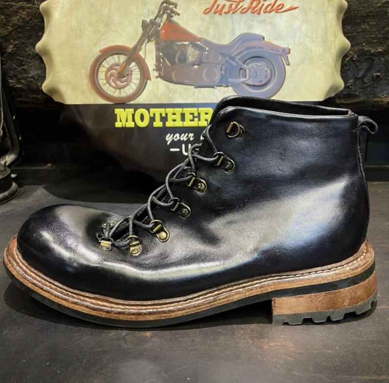 May include: Black leather boot with a rounded toe and brown sole. The boot features metal eyelets and a lace-up closure. The background includes a motorcycle graphic and the text "MOTHER your -U-".