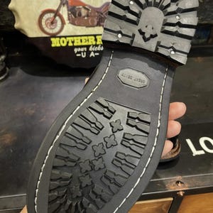 May include: Close-up of a black boot sole with a skull design on the heel. The sole has a deep tread pattern for grip and the words "GREAT DEITY" are embossed. The boot is stitched with white thread.