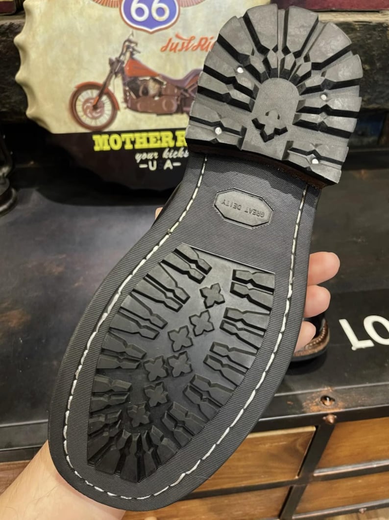 May include: Close-up of a black boot sole with a skull design on the heel. The sole has a deep tread pattern for grip and the words "GREAT DEITY" are embossed. The boot is stitched with white thread.