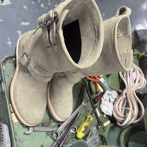 May include: A pair of light olive green suede leather boots with buckle straps. The boots have a round toe and a stacked sole. The boots are on a green surface with tools.