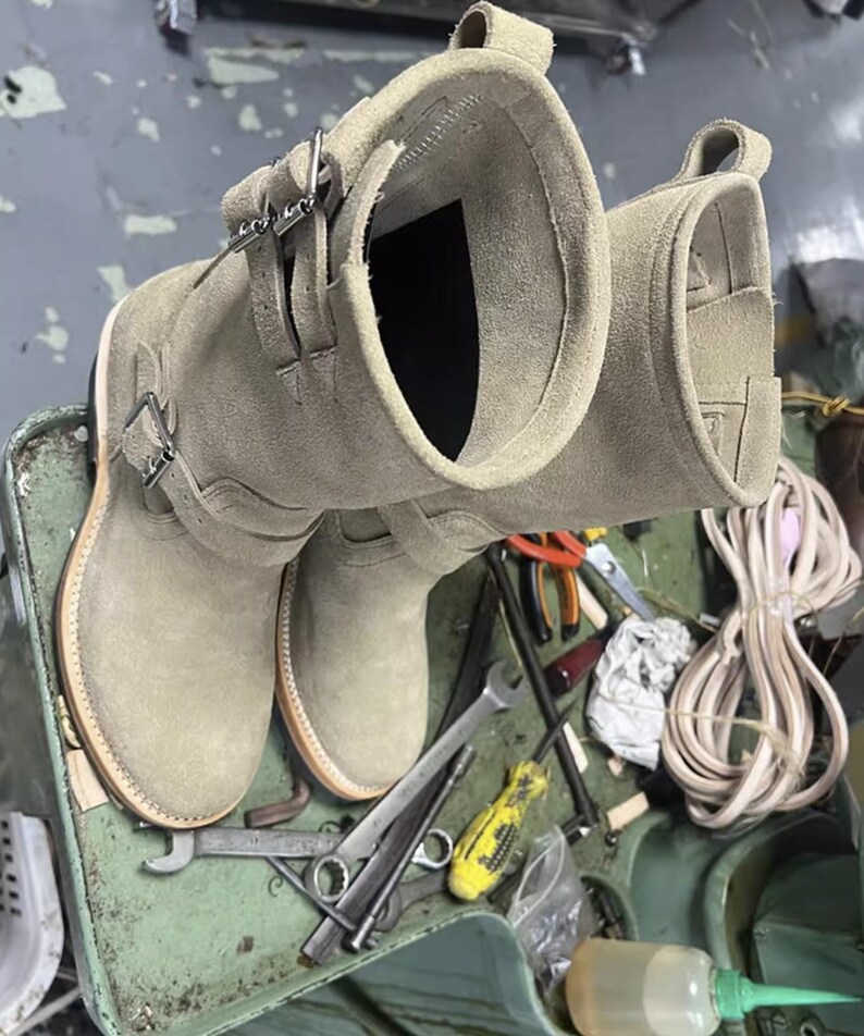 May include: A pair of light olive green suede leather boots with buckle straps. The boots have a round toe and a stacked sole. The boots are on a green surface with tools.