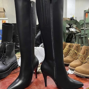 Puede incluir: Un par de botas negras hasta la rodilla con puntera afilada y tacones de aguja. Las botas tienen cierre de cremallera lateral y un acabado pulido. Los tacones miden aproximadamente 10 cm de alto. Otras botas son visibles en el fondo.