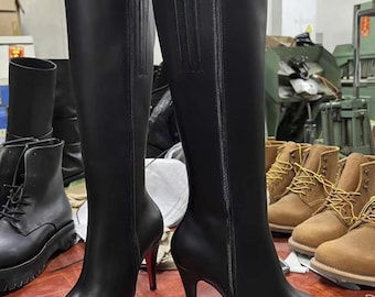 Könnte beinhalten: Ein Paar schwarze kniehohe Stiefel mit spitzer Zehenpartie und Stiletto-Absätzen. Die Stiefel haben einen Reißverschluss an der Seite und ein glattes Finish. Die Absätze sind etwa 10 cm hoch. Weitere Stiefel sind im Hintergrund zu sehen.