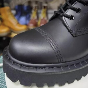 May include: Close-up of a black leather boot with a thick, lugged sole. The boot features a rounded toe with decorative stitching and black laces. The focus is on the textured details and the sturdy construction of the footwear.