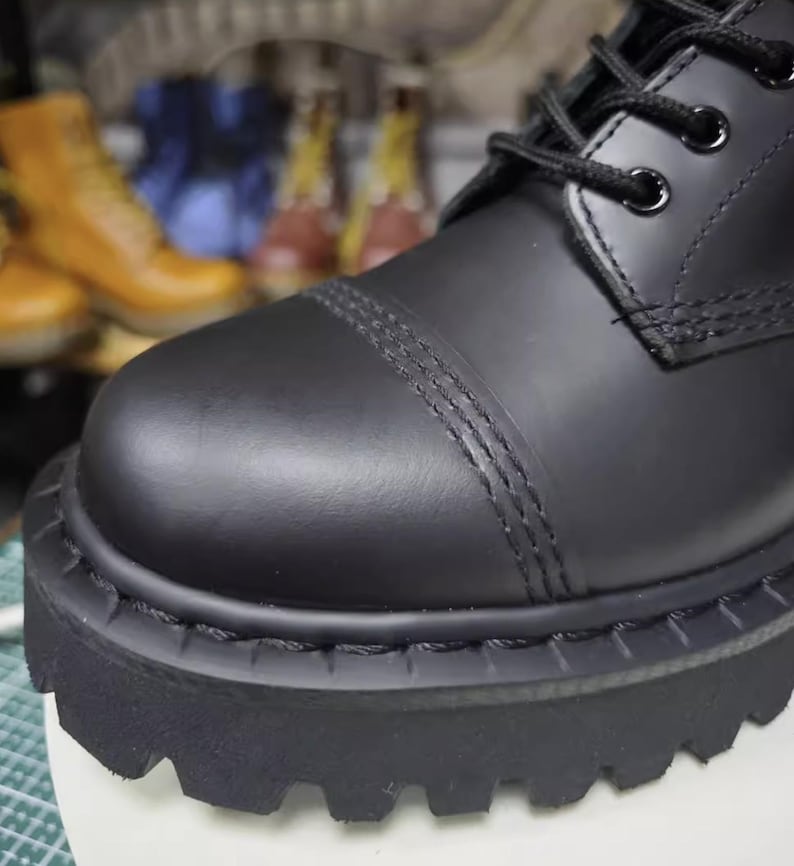 May include: Close-up of a black leather boot with a thick, lugged sole. The boot features a rounded toe with decorative stitching and black laces. The focus is on the textured details and the sturdy construction of the footwear.