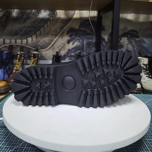 May include: Black boot sole with a deep tread pattern, designed for grip and durability. The sole features a circular indentation and a series of geometric shapes. The boot is likely part of a footwear collection.