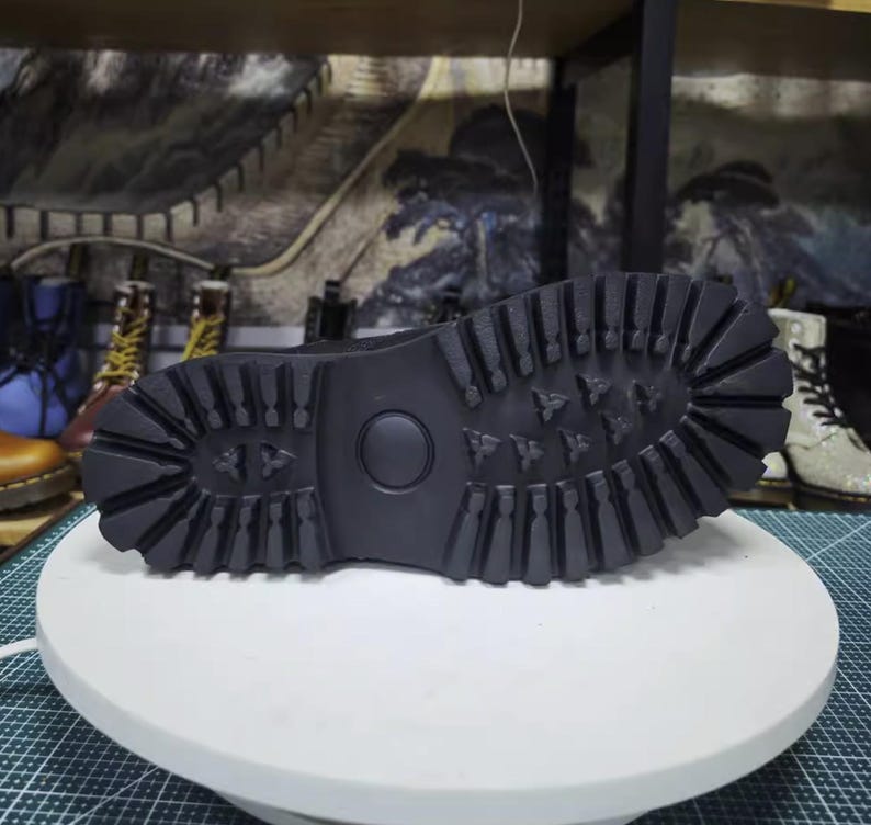 May include: Black boot sole with a deep tread pattern, designed for grip and durability. The sole features a circular indentation and a series of geometric shapes. The boot is likely part of a footwear collection.