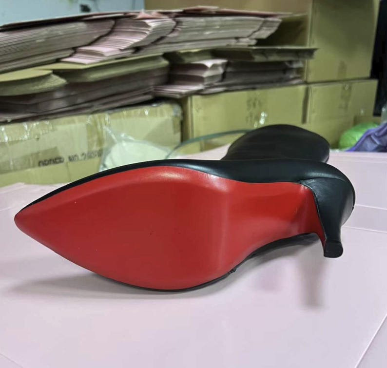 Puede incluir: Bota negra de cuero de tac&oacute;n alto con punta y suela roja. El tac&oacute;n es de estilo stiletto. La bota est&aacute; sobre una superficie rosa.