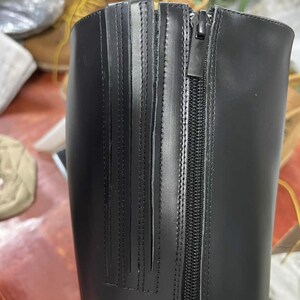 Puede incluir: Primer plano de una bota de cuero negro con cierre de cremallera. La bota presenta detalles de costura verticales en el lateral y un acabado liso y brillante. La cremallera recorre el lateral de la bota.