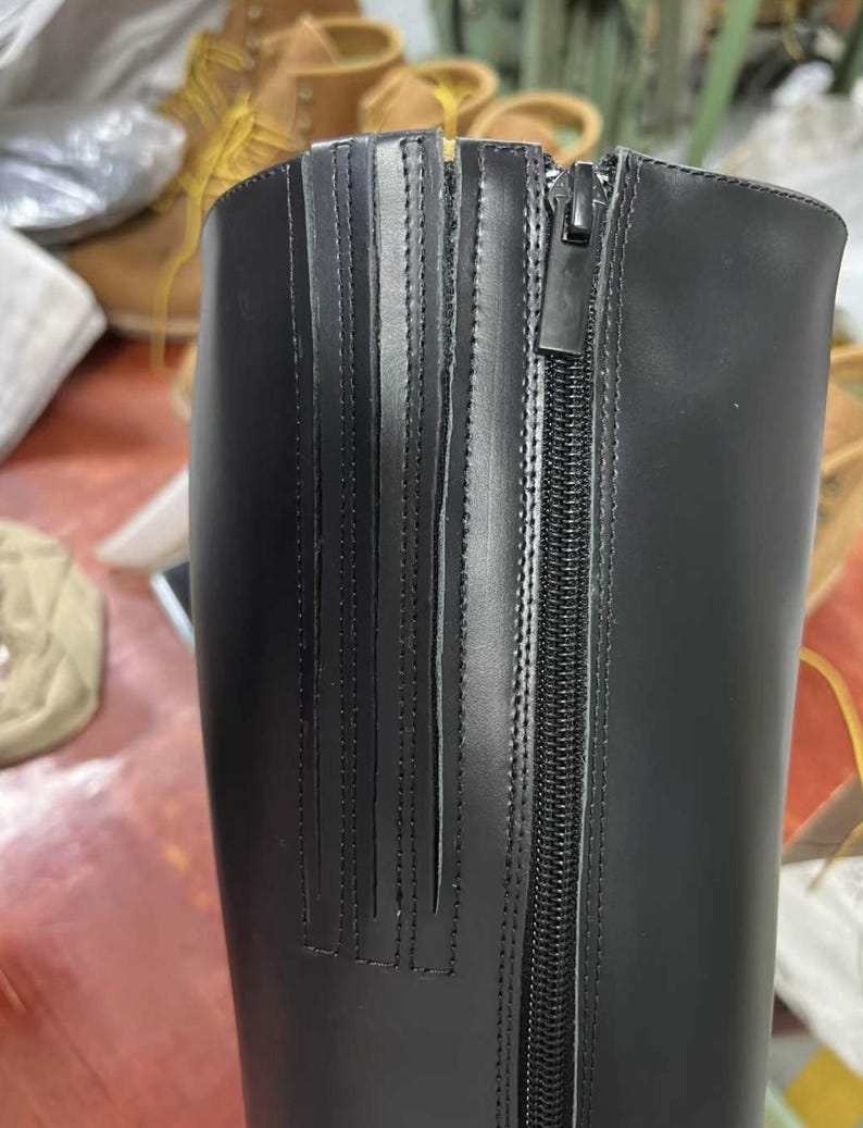 Puede incluir: Primer plano de una bota de cuero negro con cierre de cremallera. La bota presenta detalles de costura verticales en el lateral y un acabado liso y brillante. La cremallera recorre el lateral de la bota.