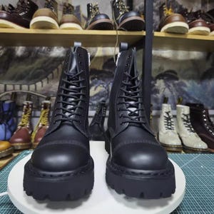 May include: A pair of black leather combat boots with thick, lug soles. The boots have a lace-up design and a zipper on the inner side. Other boots are visible on shelves in the background.