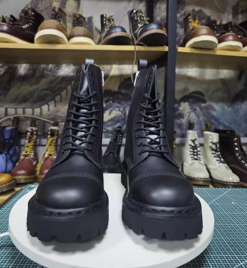 May include: A pair of black leather combat boots with thick, lug soles. The boots have a lace-up design and a zipper on the inner side. Other boots are visible on shelves in the background.