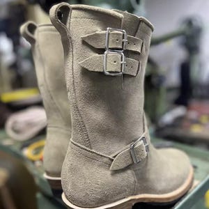 May include: Beige suede boots with silver buckles. The boots are mid-calf length and have a rounded toe. The soles are made of a light-coloured material with a dark brown base. The boots are designed in a classic, rugged style, suitable for various outfits.