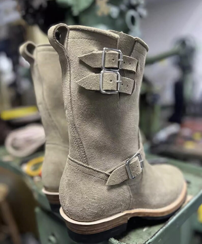 May include: Beige suede boots with silver buckles. The boots are mid-calf length and have a rounded toe. The soles are made of a light-coloured material with a dark brown base. The boots are designed in a classic, rugged style, suitable for various outfits.