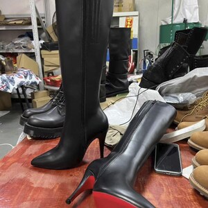 Puede incluir: Botas negras hasta la rodilla con tacones de aguja y suelas rojas. Una bota est&aacute; de pie y la otra est&aacute; tumbada. Las botas tienen cierre de cremallera y punta afilada. Otros zapatos negros son visibles en el fondo.