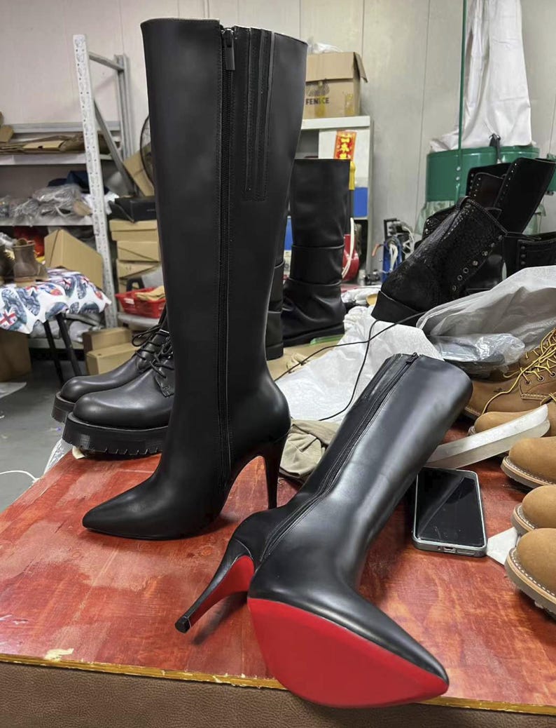 Puede incluir: Botas negras hasta la rodilla con tacones de aguja y suelas rojas. Una bota est&aacute; de pie y la otra est&aacute; tumbada. Las botas tienen cierre de cremallera y punta afilada. Otros zapatos negros son visibles en el fondo.