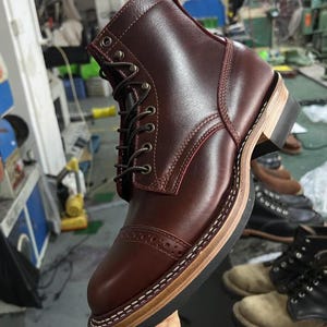 Puede incluir: Bota de cuero burdeos con puntera y cierre de cordones. La bota presenta una suela apilada con costuras visibles y una lengüeta en la parte trasera. La bota es de estilo clásico, adecuada para diversas ocasiones.