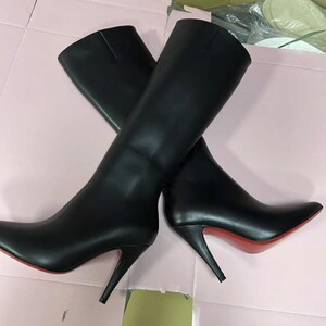 Puede incluir: Un par de botas negras hasta la rodilla con puntera y tacones de aguja. Las suelas son rojas. Las botas est&aacute;n hechas de un material liso y se exhiben sobre una superficie rosa.