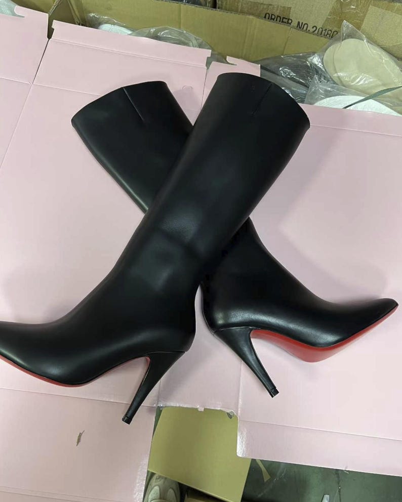 Puede incluir: Un par de botas negras hasta la rodilla con puntera y tacones de aguja. Las suelas son rojas. Las botas est&aacute;n hechas de un material liso y se exhiben sobre una superficie rosa.
