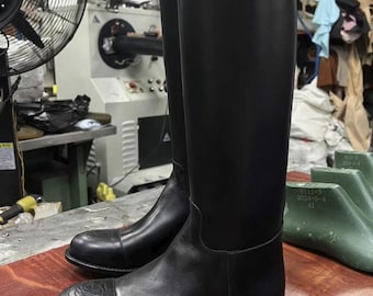 May include: A pair of tall, black leather riding boots. The boots have a smooth finish and a decorative floral design on the toe. The boots are displayed on a wooden surface, with shoemaking tools in the background.