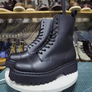 May include: A pair of black combat boots with a thick, lugged sole. The boots have a lace-up design with multiple eyelets and a rounded toe. The boots are made of a smooth, matte material and are displayed on a white surface.
