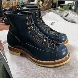 May include: A pair of navy blue leather work boots with brown laces and brass eyelets. The boots have a rounded toe, a light brown sole, and a stacked heel. The boots are sitting on a white surface.