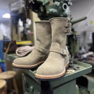 May include: Tan suede ankle boots with buckle detailing. The boots have a rounded toe and a light brown sole. The background shows a workshop setting with machinery and tools, suggesting a focus on craftsmanship and quality footwear.