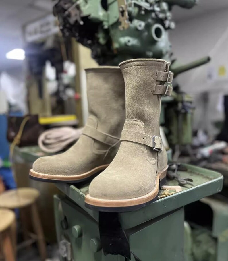 May include: Tan suede ankle boots with buckle detailing. The boots have a rounded toe and a light brown sole. The background shows a workshop setting with machinery and tools, suggesting a focus on craftsmanship and quality footwear.