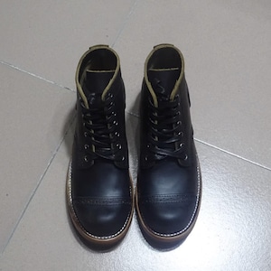 May include: A pair of black leather boots with a cap toe design and a brown rubber sole. The boots have a lace-up closure and a green stitching detail.