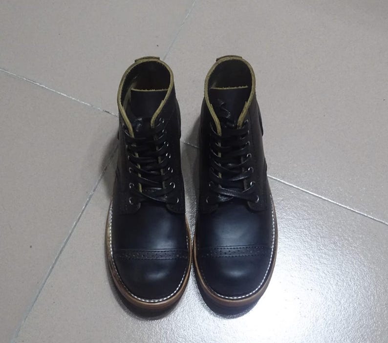 May include: A pair of black leather boots with a cap toe design and a brown rubber sole. The boots have a lace-up closure and a green stitching detail.